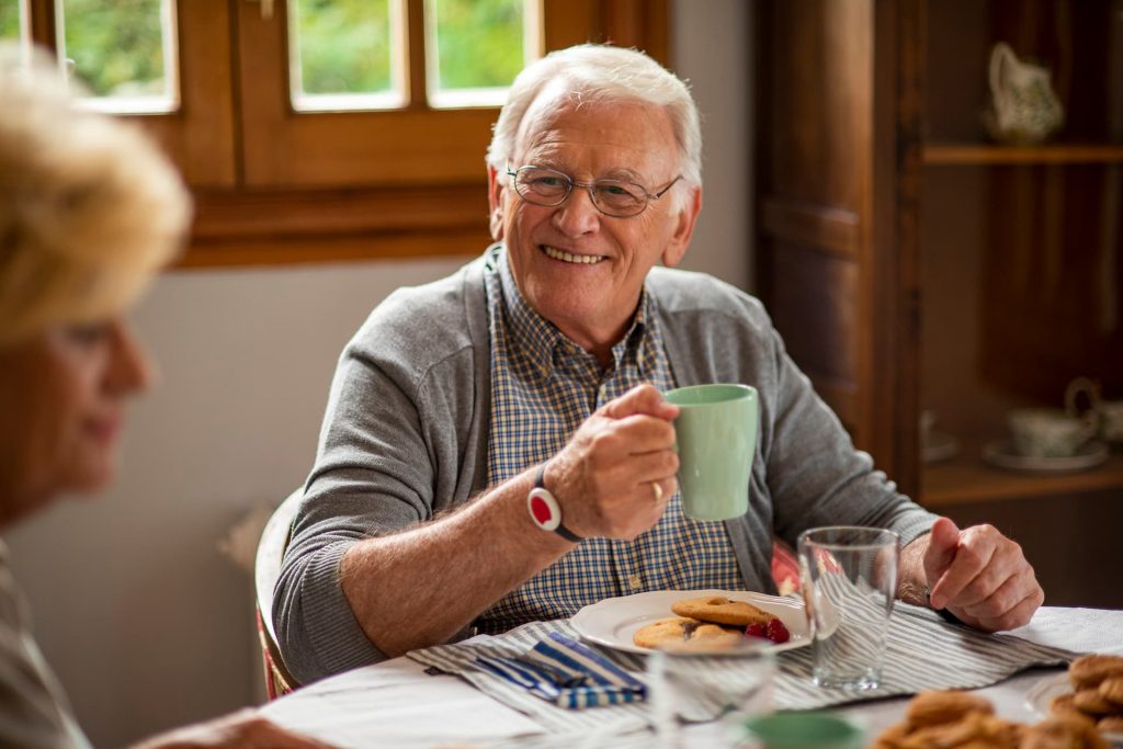 Homme senior souriant à table, portant un bracelet de téléassistance ou téléalarme pour rester en sécurité à domicile